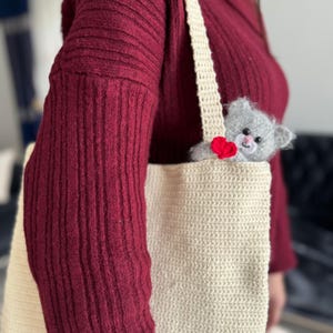 Puede incluir: Una bolsa de mano de crochet beige con un juguete de gato de crochet gris que asoma por la parte superior. El juguete de gato tiene un corazón rojo cosido en el pecho.