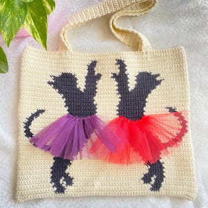 May include: A crocheted tote bag with a cream background and two black silhouettes of dogs wearing purple and red tulle tutus.