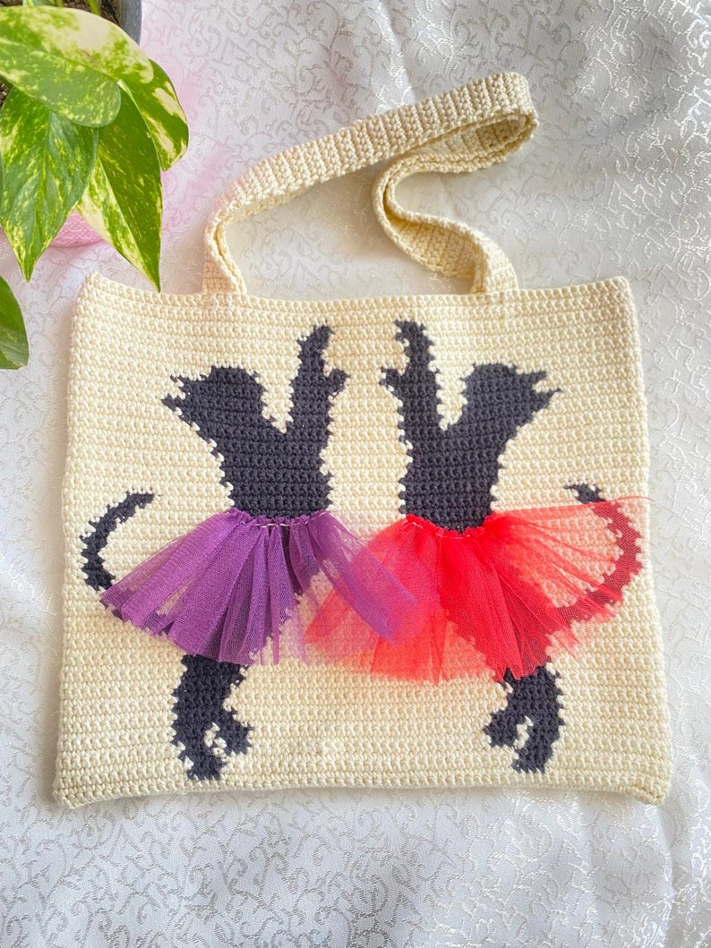 May include: A crocheted tote bag with a cream background and two black silhouettes of dogs wearing purple and red tulle tutus.
