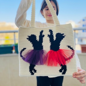 May include: A white crocheted tote bag with two black ballerina silhouettes. The ballerinas are wearing purple and red tulle tutus.