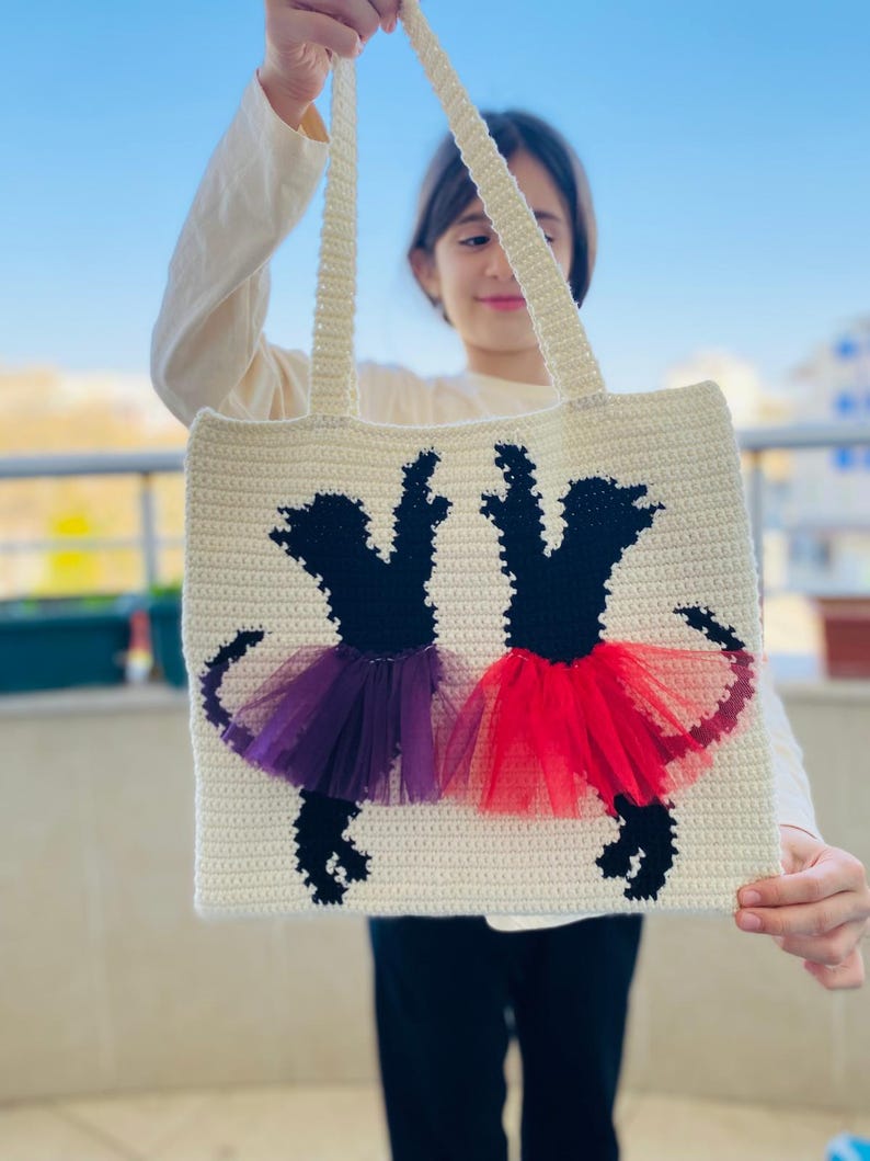 May include: A white crocheted tote bag with two black ballerina silhouettes. The ballerinas are wearing purple and red tulle tutus.