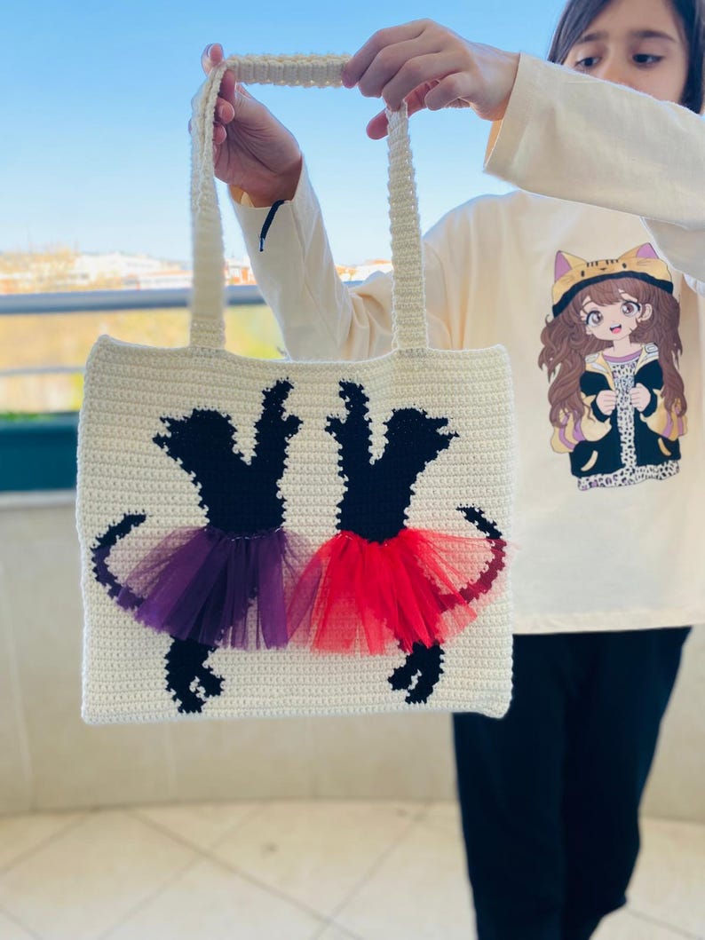May include: A white crocheted tote bag with black silhouettes of two dancers wearing purple and red tulle tutus.