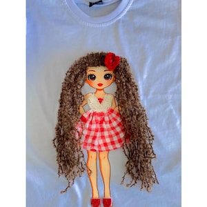 Puede incluir: Una camiseta blanca con el gráfico de una muñeca que tiene el pelo largo de lana marrón texturizada, un accesorio de flor de tela roja y un vestido de cuadros vichy rojo y blanco con corpiño de encaje blanco, zapatos rojos y rostro impreso.