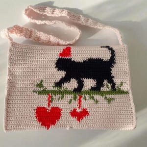 May include: A light pink crocheted shoulder bag. It features a black cat wearing a red hat, sitting on a green branch with two red hearts hanging below. The strap is also crocheted in a matching pink colour.