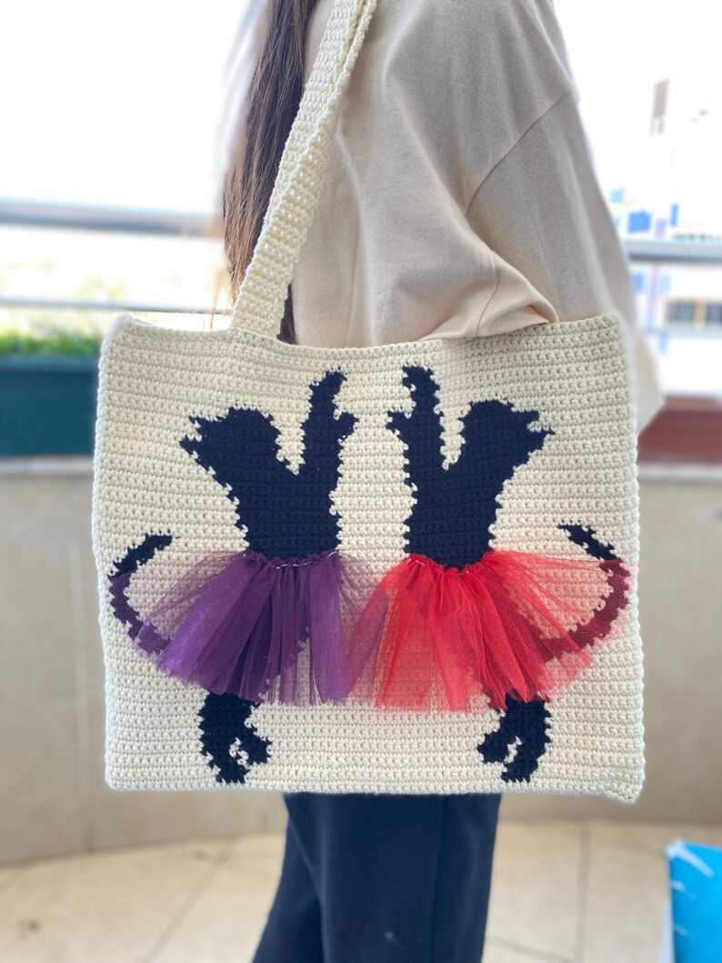 May include: A white crocheted tote bag with black silhouettes of two dogs wearing purple and red tulle tutus.