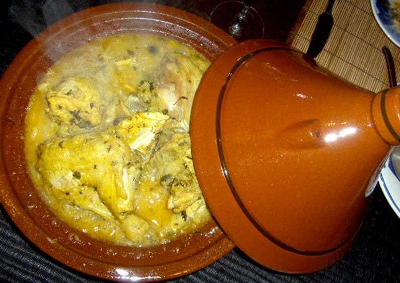 May include: A brown clay tagine pot with a lid, filled with a chicken stew. The stew is a light yellow color and has a few green herbs in it. Steam is rising from the pot.