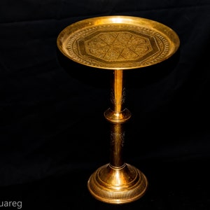 May include: A gold metal decorative stand with a round top and a detailed engraved design. The stand has a tall, cylindrical base.