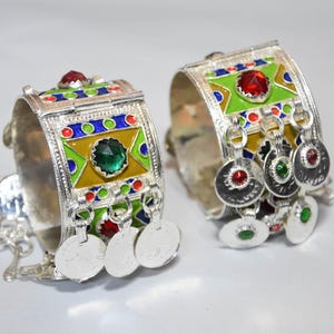 May include: A pair of silver-toned metal cuff bracelets featuring vibrant enamel inlays of lime green, blue, and yellow. Each bracelet is adorned with faceted red and green glass stones and dangling circular charms with small colored accents.