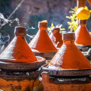 May include: A row of traditional Moroccan tagines, with terracotta lids and bases, are steaming on a stovetop. The tagines are arranged in a line, with the lids slightly ajar, revealing the contents within.