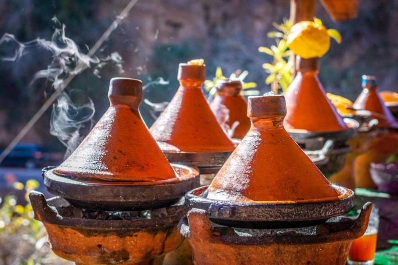 May include: A row of traditional Moroccan tagines, with terracotta lids and bases, are steaming on a stovetop. The tagines are arranged in a line, with the lids slightly ajar, revealing the contents within.