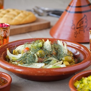 May include: A close-up of a traditional Moroccan tagine dish with fennel, olives, and other vegetables. The dish is served in a terracotta tagine pot with a conical lid. The tagine is surrounded by small bowls of sauce and rice.