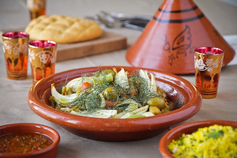 May include: A close-up of a traditional Moroccan tagine dish with fennel, olives, and other vegetables. The dish is served in a terracotta tagine pot with a conical lid. The tagine is surrounded by small bowls of sauce and rice.
