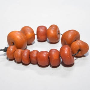 May include: A strand of eleven smooth, rounded, amber-colored beads of varying sizes strung on a thin black cord. The beads have a polished, semi-translucent appearance with irregular, organic shapes and a warm, earthy orange-brown hue.