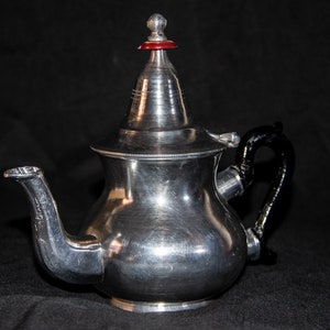 Puede incluir: Una tetera de plata con asa negra y tapa roja y plateada. La tetera tiene un cuerpo redondeado y un pico en el lateral.