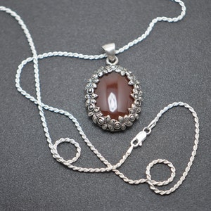 May include: A silver chain necklace with a brown oval gemstone pendant. The pendant is surrounded by a silver frame with floral details.