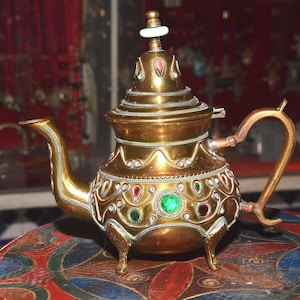 Könnte beinhalten: Eine goldene Teekanne mit aufwendigen Details und grünen und roten Edelsteinen. Die Teekanne hat einen runden Körper und einen geschwungenen Ausguss. Sie steht auf einer bunten Oberfläche.