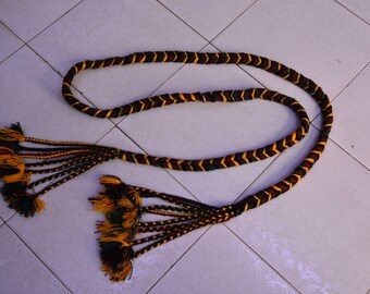 May include: A long, braided cord with black, yellow, and green stripes. The cord has tassels on both ends, each with multiple strands of black, yellow, and green.