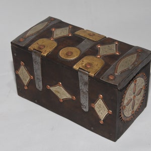 May include: A small wooden box with intricate metalwork. The box is decorated with brass and silver accents in a geometric pattern. The box has a hinged lid and a decorative metal clasp.