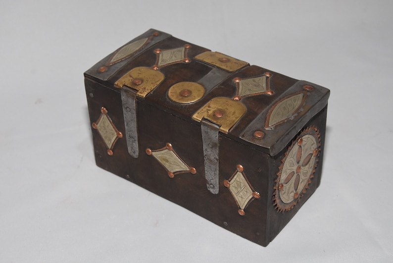 May include: A small wooden box with intricate metalwork. The box is decorated with brass and silver accents in a geometric pattern. The box has a hinged lid and a decorative metal clasp.