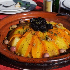 May include: A close-up of a traditional Moroccan tagine dish with yellow and orange vegetables, olives, and a layer of dried prunes on top. The tagine is served in a brown clay pot.