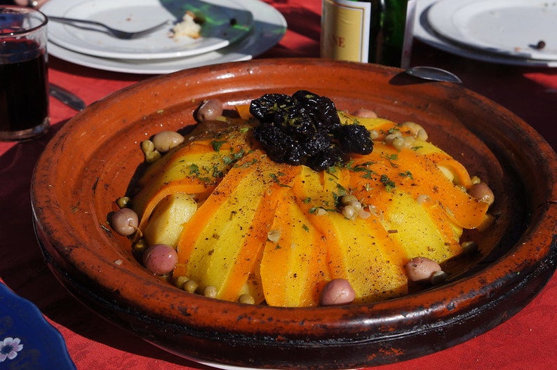 May include: A close-up of a traditional Moroccan tagine dish with yellow and orange vegetables, olives, and a layer of dried prunes on top. The tagine is served in a brown clay pot.