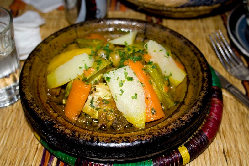 May include: A close-up of a traditional Moroccan tagine dish, featuring a brown clay pot filled with a stew of vegetables and meat. The stew includes carrots, potatoes, green beans, and herbs, all simmered in a flavorful broth.