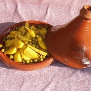 May include: A terracotta tagine with a lid, filled with yellow potatoes and green olives. The tagine is sitting on a purple tablecloth.