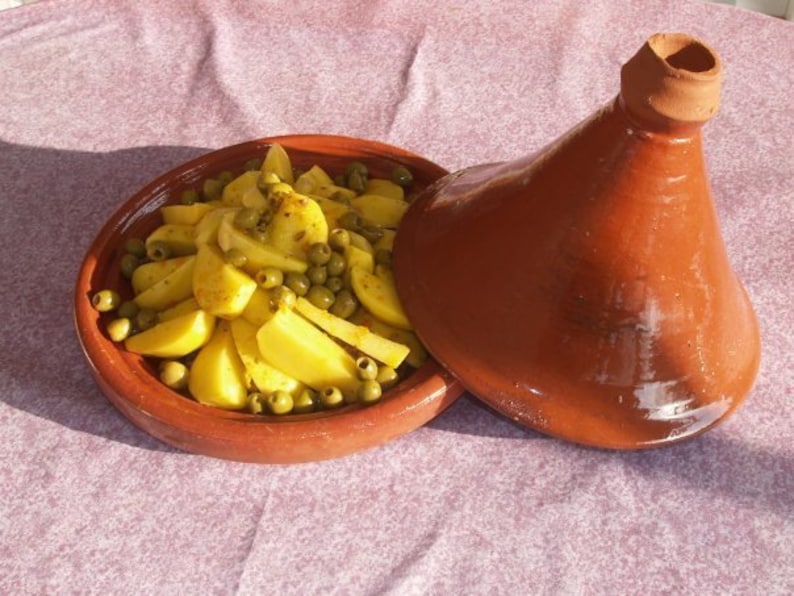 May include: A terracotta tagine with a lid, filled with yellow potatoes and green olives. The tagine is sitting on a purple tablecloth.