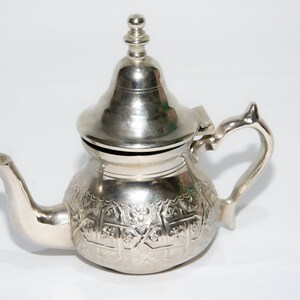 Puede incluir: Una tetera de plata con un diseño ornamentado y una tapa con un pomo. La tetera tiene un pico y una asa.