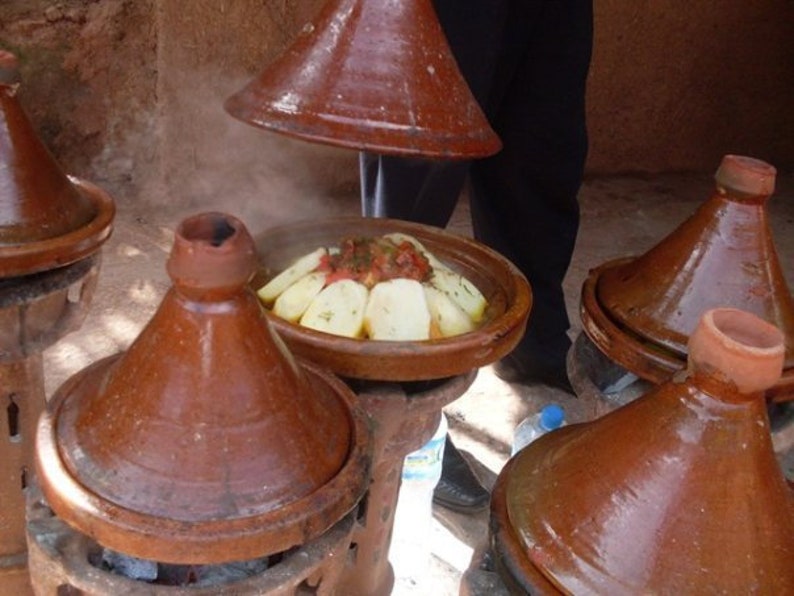 May include: A close-up of a traditional Moroccan tagine cooking on a stovetop. The tagine is made of brown clay and has a conical lid. The food inside the tagine is steaming and appears to be a stew with vegetables and meat.