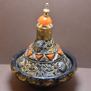 May include: A black and gold Moroccan tagine with an orange lid. The tagine is decorated with intricate floral patterns and has a round base.