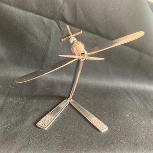 May include: A handcrafted metal sculpture of an aeroplane, constructed from repurposed silverware. The wings are formed from knives, the body from a light bulb base, and the tail from a fork. The sculpture stands on a base of two spoons.