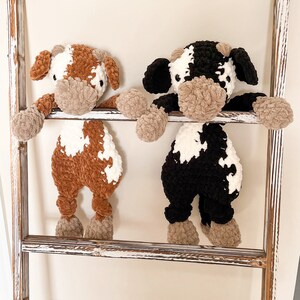 May include: Two crocheted stuffed cows, one brown and white and one black and white, are hanging on a whitewashed wooden ladder.