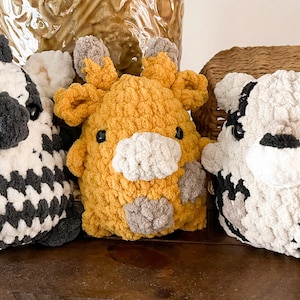 May include: Three crocheted stuffed animals, a zebra, a giraffe, and a cow, are sitting on a wooden surface. The zebra is black and white, the giraffe is yellow and brown, and the cow is white and black.