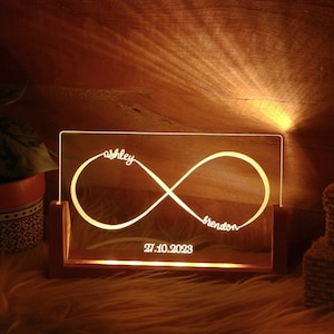 Può includere: Un cartello luminoso in legno con un simbolo di infinito e i nomi "Ashley" e "Brendon" incisi. La data "27.10.2023" è anche inclusa sul cartello.