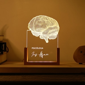 May include: A brain-shaped lamp with a wooden base, illuminated to show the brain's structure. The lamp has the words "PSICÓLOGA" and "Iris Alfaro" etched on the clear acrylic panel. The light emits a warm glow.