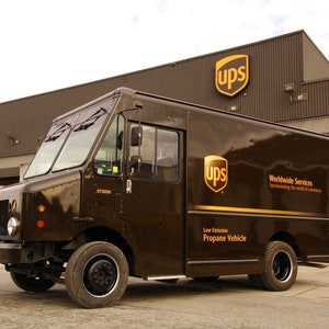 May include: A brown UPS delivery truck with the company logo and "Worldwide Services" text on the side. The truck is parked in front of a brown building with the UPS logo on the side.