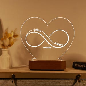 Peut inclure: Une veilleuse en acrylique blanche en forme de cœur avec un symbole d'infini et les noms "jessica" et "jackson" inscrits. La date "28.08.2023" est également inscrite sur la lumière.