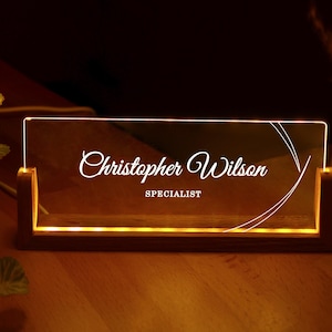 Könnte beinhalten: Beleuchtetes Namensschild mit dem Namen "Christopher Wilson" und dem Titel "Specialist" in weißer Schrift. Das Namensschild aus klarem Acrylglas steht in einem Holzsockel mit warmer Beleuchtung.