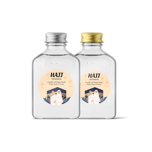 May include: Two clear glass bottles with silver and gold lids. Each bottle has a round label with a light orange background and a graphic of two people in white robes standing in front of a blue and gold building. The text on the label reads "Hajj Mubarak, A gift of Zam Zam, From Name & Name".