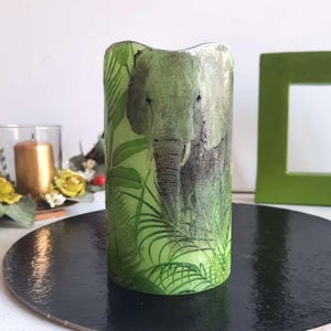 以下が含まれることがあります： 白い象と緑の葉のデザインが施された緑色のろうそく。ろうそくは黒い表面の上に置かれています。