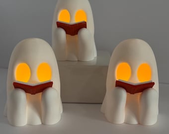 May include: Three white ghost-shaped night lights, each holding a red book. The ghosts have glowing orange eyes and are sitting down. The lights are on a white surface, perfect for Halloween or spooky decor.