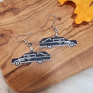 May include: A pair of silver dangle earrings with black and white car charms. The cars are shaped like hearses with a white trim and a black interior.