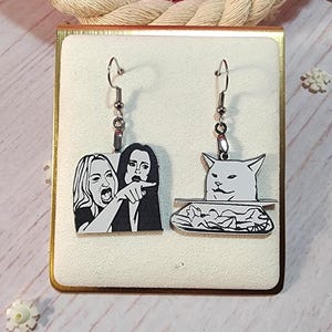 May include: Silver-tone dangle earrings featuring two meme designs. One earring depicts the woman yelling at a cat meme, and the other shows the cat meme. The earrings are suspended from silver-colored fish hook ear wires.