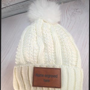 May include: Cream-colored knit beanie with a fluffy white pom-pom on top. The hat features a ribbed knit design and a brown leather patch with the text "Name engraved here".