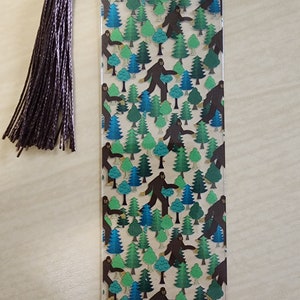 May include: A clear plastic bookmark with a brown tassel. The bookmark features a repeating pattern of brown Bigfoot figures walking through a forest of green and blue trees.