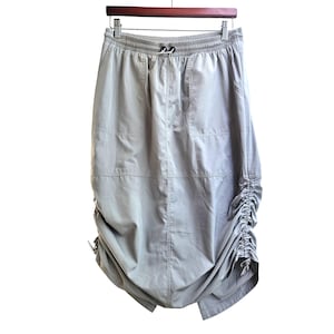 Könnte beinhalten: Hellgrauer Rock mit elastischem Bund und Kordelzug. Der Rock hat aufgesetzte Taschen und geraffte Details an den Seiten, wodurch eine einzigartige Silhouette entsteht. Er hängt an einem Holzkleiderbügel.