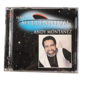 Puede incluir: Una caja de CD con el álbum "Serie Millennium 21" de Andy Montañez. La portada tiene un diseño azul y negro con temática espacial con la foto del artista en un marco blanco. La caja del CD es de plástico transparente.