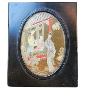 May include: A framed oval artwork featuring a traditional Asian scene. The artwork depicts three figures in colorful attire, with a balcony and foliage in the background. The frame is a dark, rectangular shape.