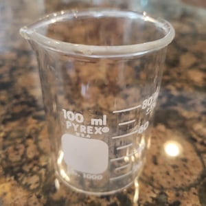 May include: A clear glass beaker with a white label that reads "100 ml PYREX USA". The beaker has markings for measuring liquids in milliliters.